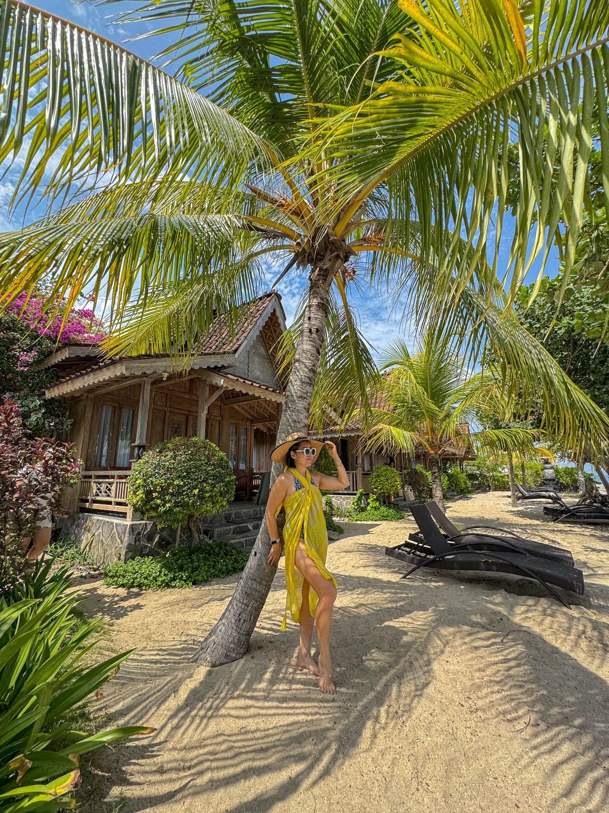 Woman in a yellow sarong leaning on a palm tree beside wooden beach huts