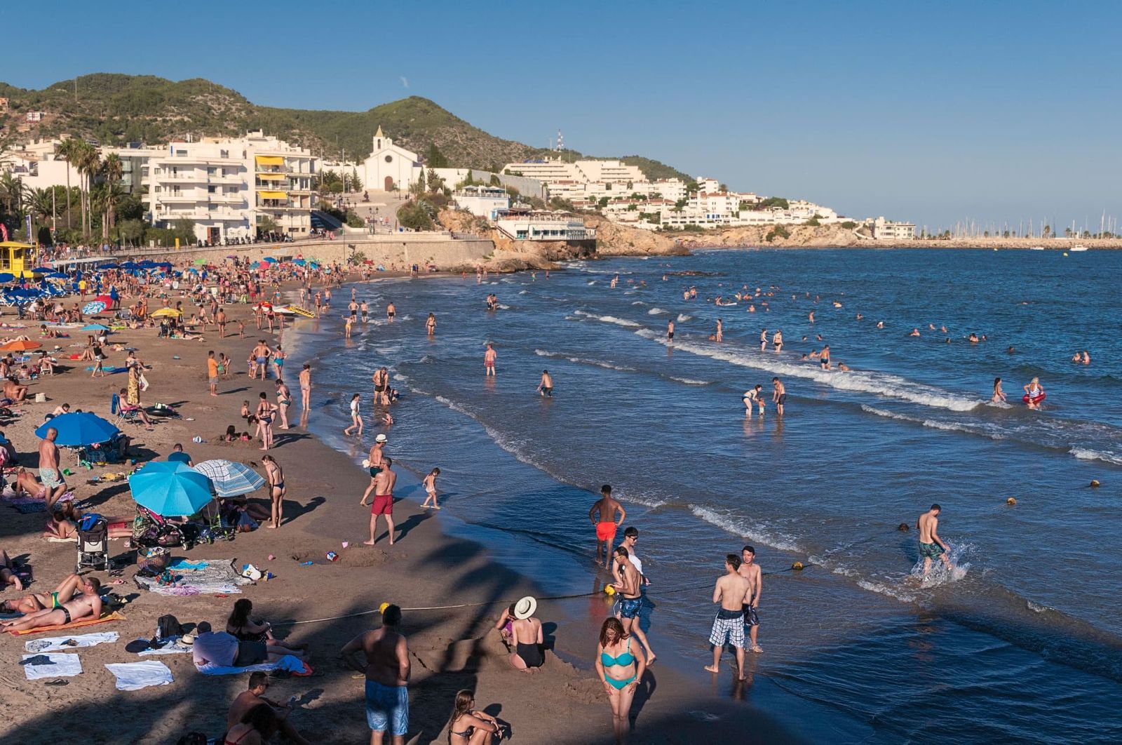 Busy beach below white coastal buildings and hills