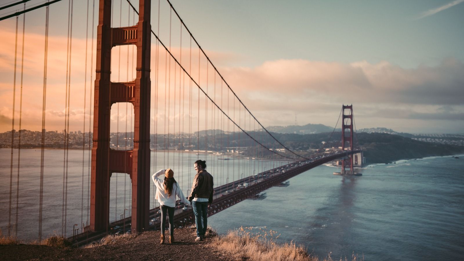 Couple holding hands on a hill looking at the Golden Gate Bridge at sunset
